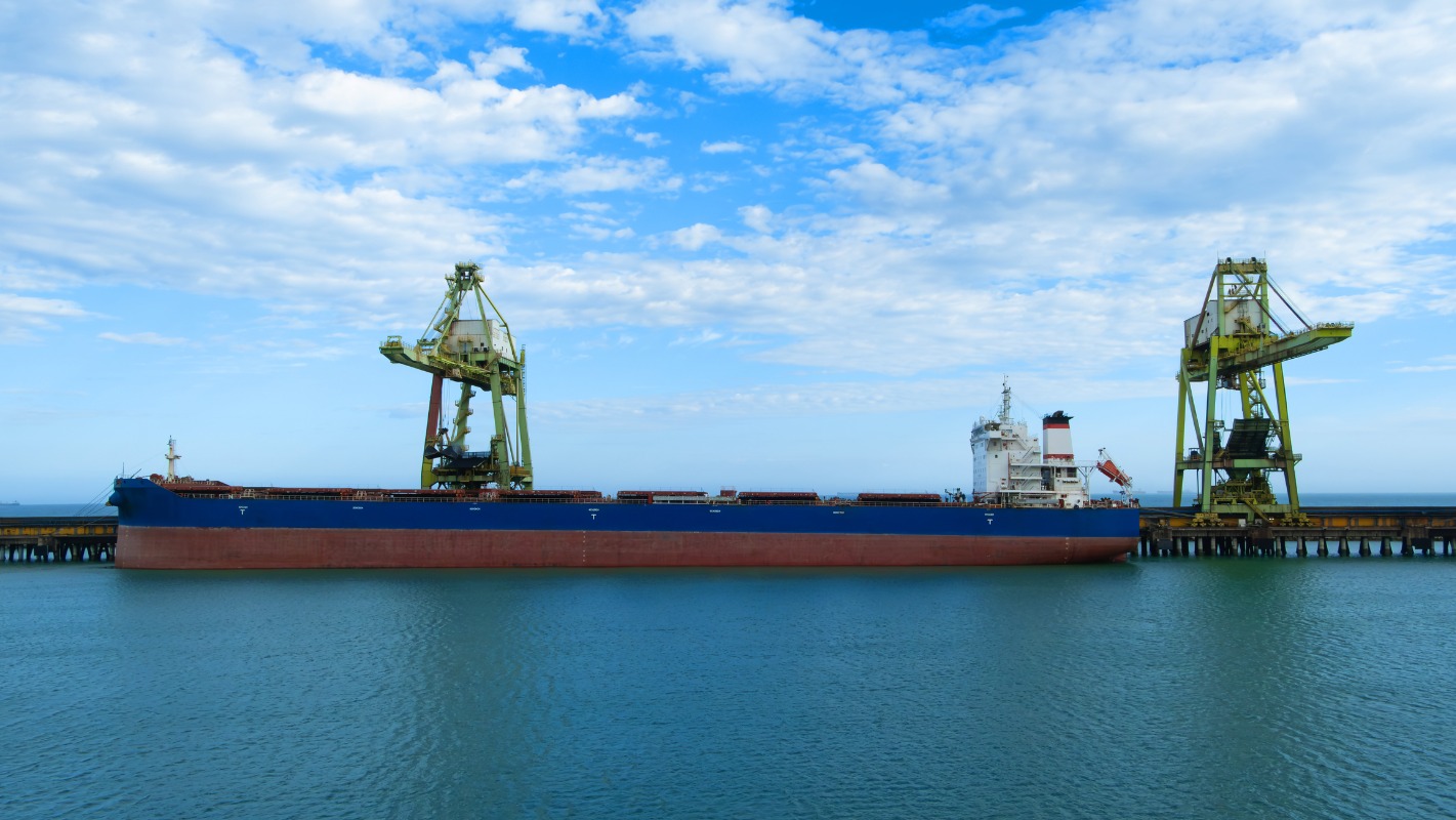 Blue freight ship is docked between two large industrial cranes at a pier